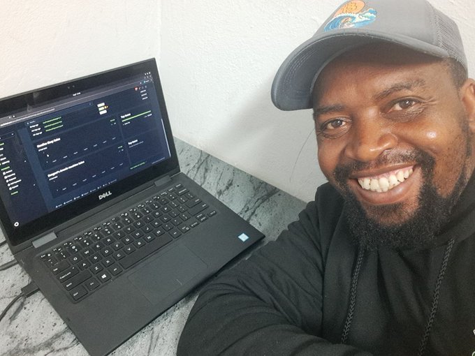 A smiling man wearing a gray Miami Dolphins cap and black hoodie sits next to a Dell laptop displaying what appears to be a financial or data dashboard.