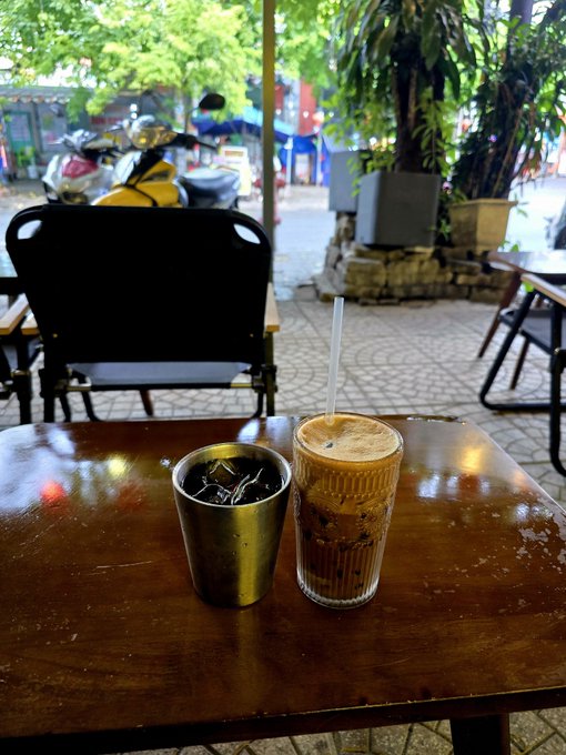 A glass of iced coffee sits on a wooden table beside a metal cup, with a leaf-shaded street scene and parked motorbikes visible in the background. The setting captures the post's motivational spirit of fueling up with coffee to stay sharp and keep building, one day at a time.