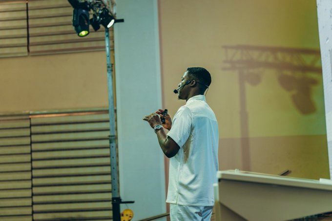 A man wearing a headset microphone and white shirt gestures while speaking on stage, with a projection screen behind him displaying a bridge silhouette. The image captures a public speaking moment that visually reinforces the post's message about staying current with trending topics and adapting to change.