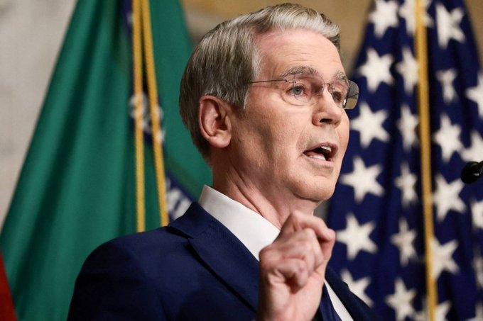 A man with silver hair and glasses, wearing a dark suit, speaks emphatically while gesturing with his hand, with flags visible in the background. The image appears to relate to the G20 Finance Ministers meeting mentioned in the post, though the man pictured does not appear to be Fed Chair Kevin Warsh or Treasury Secretary Scott Bessent based on visible features.