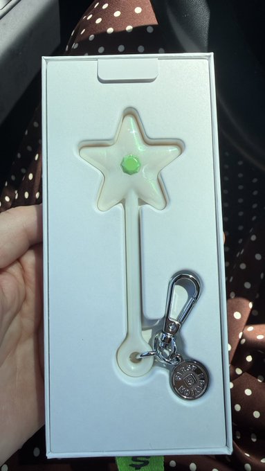 The image shows a person holding up a white keychain with a green gemstone star design, which appears to be made of plastic or clear material. The keychain has a silver ring attached to it, possibly for attaching keys or other small items. The background is blurred and does not provide any additional information about the context of the image.