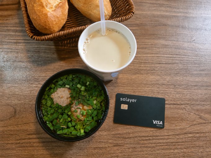 A wooden table holds a bowl of soup with green onions, a cup of milk tea, a basket of bread, and a black 'solayer' Visa Platinum card.