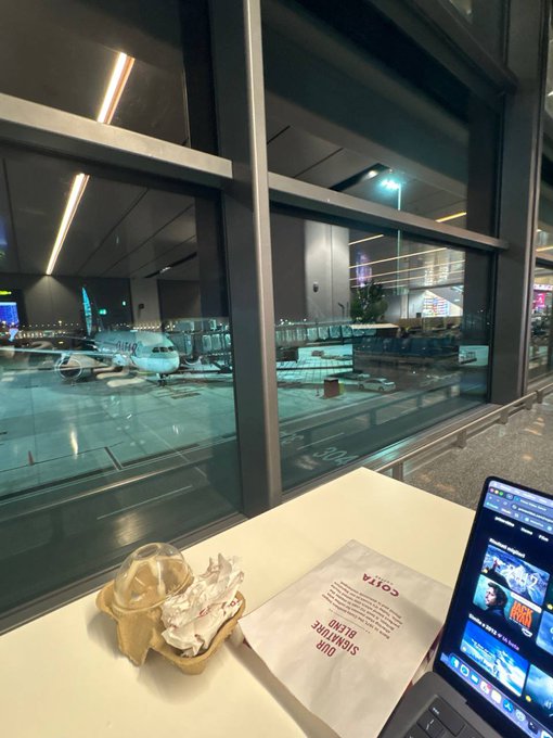 A view from an airport gate shows a Qatar airplane on the tarmac, with a laptop displaying a streaming service and a food container on a tray table in the foreground.
