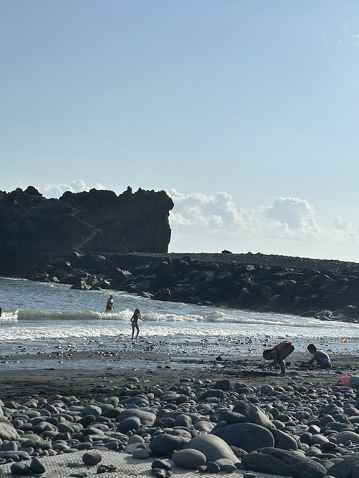 A scenic beach view shows people wading and playing near the shoreline, with a rocky breakwater and cliffs visible in the background under a partly cloudy sky.
