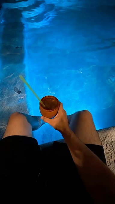 A first-person view of a hand holding a clay cup with a green straw, dangling legs over the edge of a bright blue swimming pool at night.