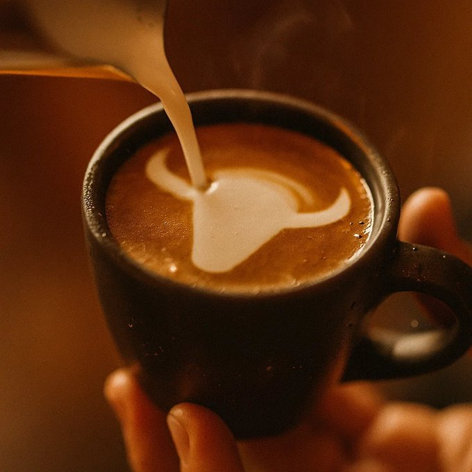 A close-up of milk being poured into a dark ceramic cup of coffee, creating latte art in warm, ambient lighting.