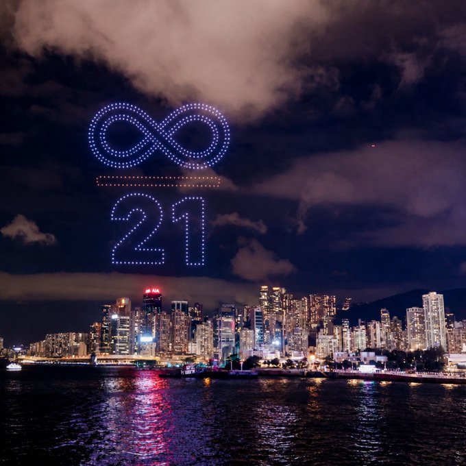 A nighttime cityscape over water features a drone light show forming an infinity symbol above the glowing number '21' against the cloudy sky.