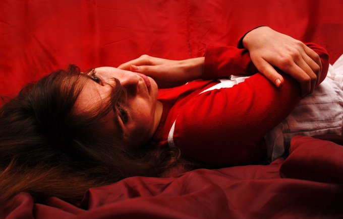 A young woman with dark hair lies on red bedding, wearing a red sweater, looking upward with a thoughtful expression as she rests on white pillows.
