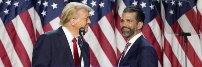 Donald Trump and Donald Trump Jr. stand smiling together on a stage with American flags in the background.