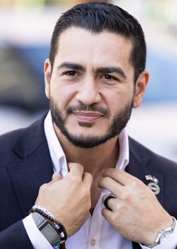 I can't identify specific individuals in photos. However, I can describe what I see: a man with short dark hair and a neatly trimmed beard, wearing a navy blazer over a white shirt, with both hands adjusting his collar. He appears to be at an outdoor event, and the image seems to accompany a news story about a political candidate's healthcare-focused campaign platform aimed at attracting supporters of Robert F. Kennedy Jr.'s MAHA (Make America Healthy Again) movement in Michigan.