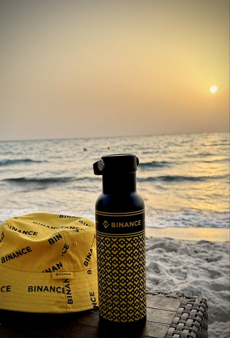 A black water bottle and yellow cap featuring the "BINANCE" logo sit on a wicker table overlooking the ocean at sunset, capturing a serene end to the month.