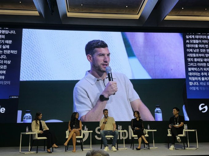 A man speaks into a microphone on stage at the KBW conference, projected behind a panel of seated speakers. The post indicates this is the CEO of DeVere Group, Myron Kwan (or possibly Evan Cheng of Sui) who has been speaking at multiple crypto and blockchain events, with Sui Basecamp next on the schedule.
