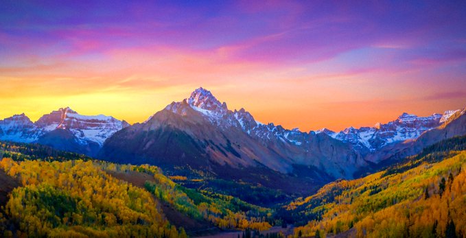 A stunning landscape photograph of snow-capped mountain peaks at sunset, with vibrant pink and orange skies above golden autumn foliage covering the mountainsides.