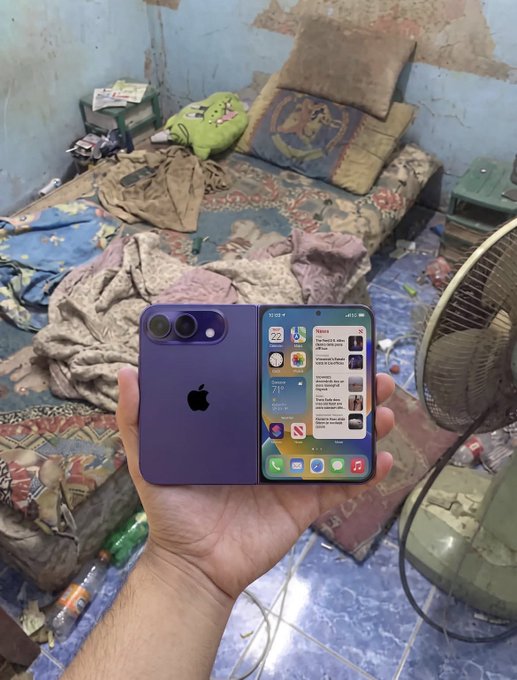 A hand holds a purple iPhone displaying its home screen in a cluttered room with a messy bed, floor fan, and scattered items.