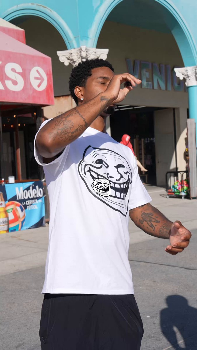 The image shows a man wearing a white t-shirt with an extremely funny face on it, which appears to be a troll from Venice edition. The man has a tattoo of a cat on his arm and he's standing next to a street with some stores visible in the background. There is also a red banner that says "Troll The World: Venice Edition". This gives context to the content of the image as it seems related to the post, which features a funny, troll-themed design or illustration.