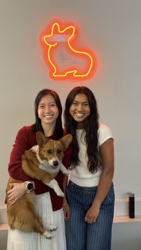 Two smiling women pose with a corgi in front of a glowing neon dog sign, capturing a fun moment at the Corgi office during the Willow voice-maxxing challenge event.