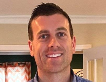 A man with short brown hair and a bright smile, wearing a blue collared shirt, stands indoors in front of a doorway with patterned curtains. The image accompanies a post featuring Chainlink Labs' Andrew McCormick, likely depicting him as he discusses AI agents and robots using digital assets and blockchains for transactions at the SALT conference.