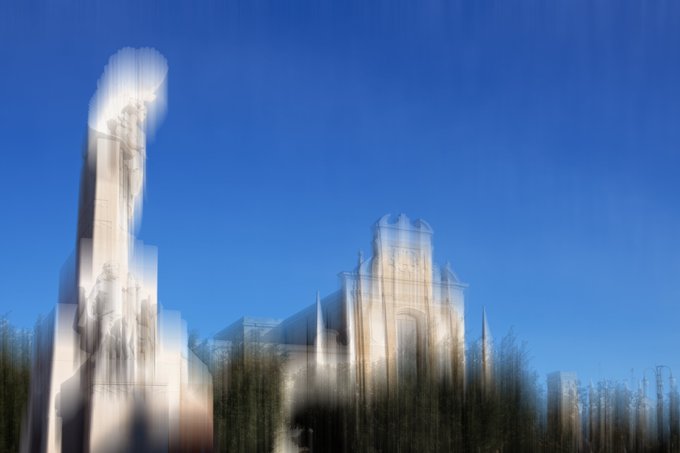 A motion-blurred photograph showing what appears to be a white cathedral or church building against a bright blue sky, with trees in the foreground.