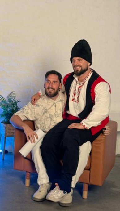 Two men in traditional Balkan-style clothing pose together on a brown leather chair, one wearing a patterned shirt holding a rolled paper and the other in a red and black vest with a tall black hat.