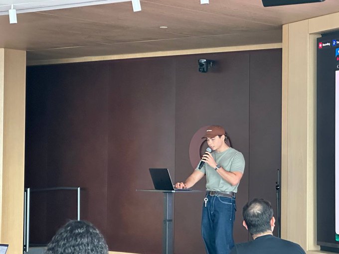 The image shows a man standing at a podium, giving a presentation or speech while holding a microphone. The room appears to be a conference room with a brown wall behind him. There are several people sitting around the room, possibly listening to his presentation or waiting for their turn to speak. One person is wearing a baseball cap and has a laptop on a table in front of them.
