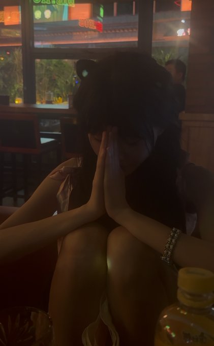In a dimly lit restaurant with a window showing the storefront "SUSHI RAN" and "SUSHI" signage, a woman wearing cat ears sits with her hands pressed together in a prayer-like gesture, accompanied by a water bottle on the table.