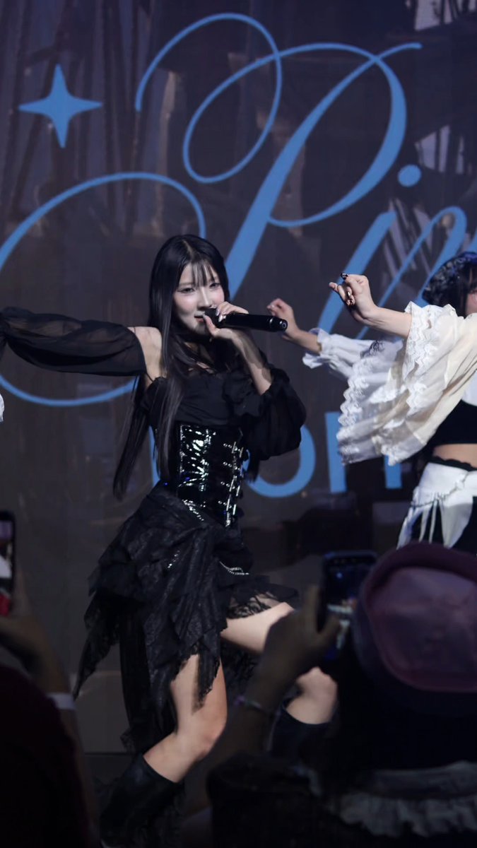 A female performer in a black dress with a corset-style bodice sings into a microphone on stage, with an elaborate 'IVE' logo displayed on the backdrop.