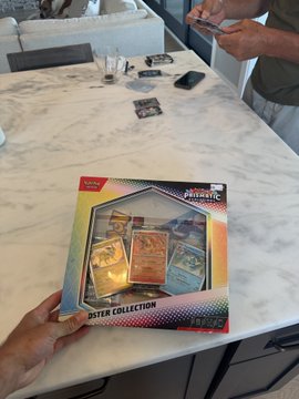 A hand holds a 'Pokémon TCG: Surging Sparks Elite Trainer Box' on a marble countertop while another person uses a phone in the background.