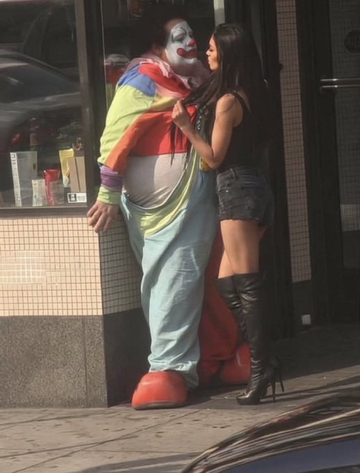 A street photograph showing a person in a fat clown costume interacting closely with a woman wearing a black top, denim skirt, and high black boots against a tiled wall.