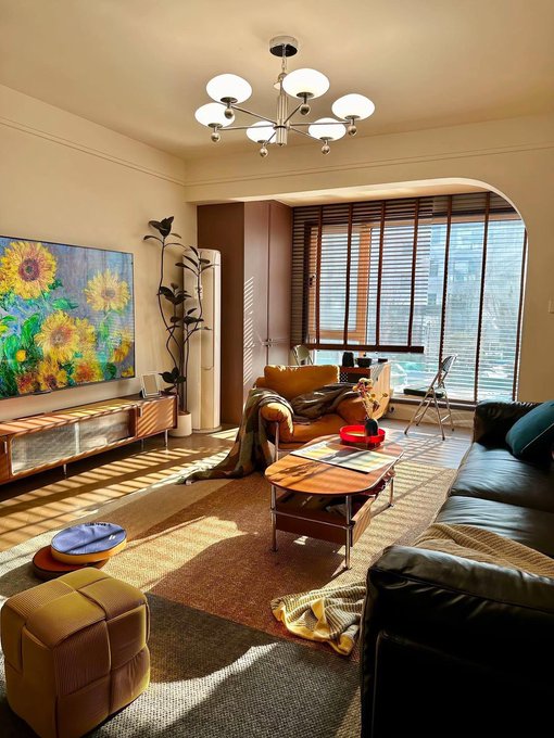A photograph of a modern, cozy living room with a large TV displaying sunflowers, a black leather sofa, a yellow ottoman, a mid-century chandelier, and warm afternoon sunlight streaming through window blinds.