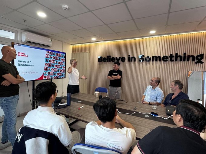 An indoor meeting room scene where a group of people are gathered for a presentation; a screen displays 'Investor Readiness' with logos for TMC and ANGELS, and a wall sign reads 'Believe in something'.