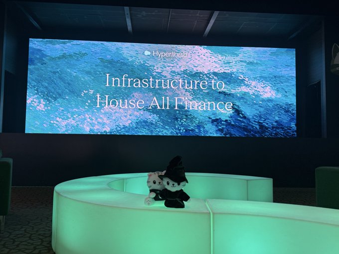 A display screen at a Hyperliquid event reading 'Infrastructure to House All Finance,' with a plush toy sitting on a glowing green curved couch below.