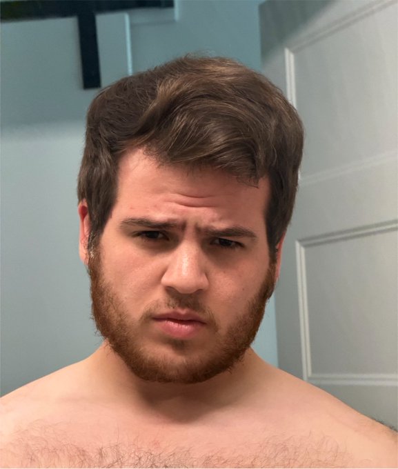 A shirtless man with brown hair and a beard looks into the camera with a slightly confused or displeased expression in what appears to be a bathroom.