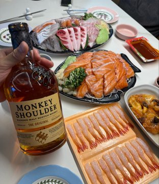 A dinner spread featuring a bottle of Monkey Shoulder whiskey alongside plates of sashimi (salmon and tuna), sushi, and other dishes on a table.