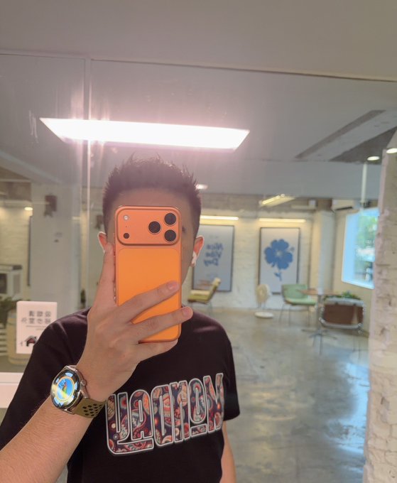A mirror selfie of a young man taking a photo with an orange iPhone, wearing a black t-shirt with a graphic print and a watch, with AirPods in his ears.