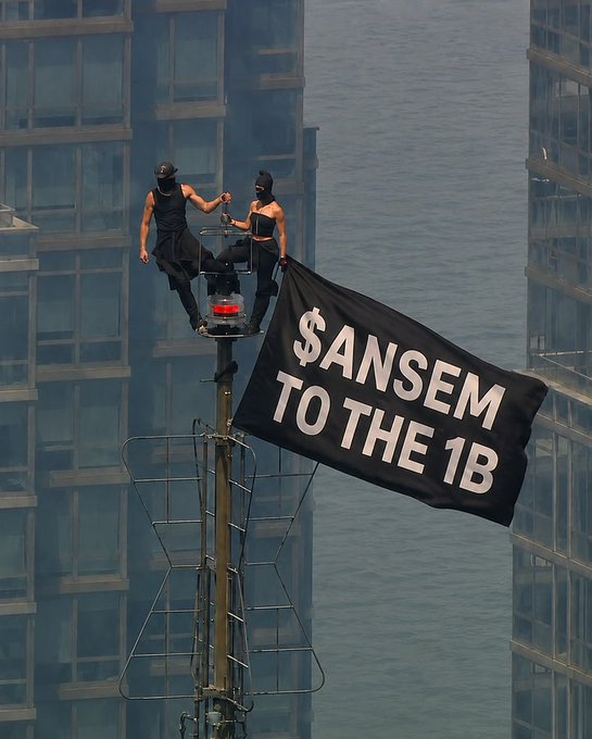 Two figures dressed in black standing atop an antenna or tower high above city buildings, holding a black banner reading '$ANSEM TO THE 1B'.
