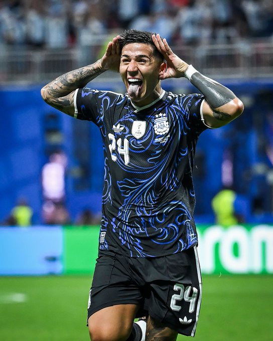 Soccer player wearing jersey number 24 celebrates with hands to his head and tongue out, wearing a dark blue and black patterned Argentina AFA kit.