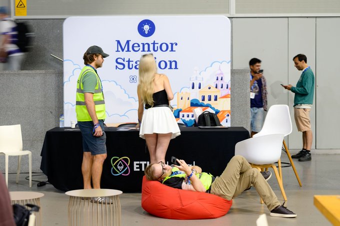 People interact at a 'Mentor Station' booth featuring ETH branding, with attendees including a man lying on a red beanbag chair and others standing around.