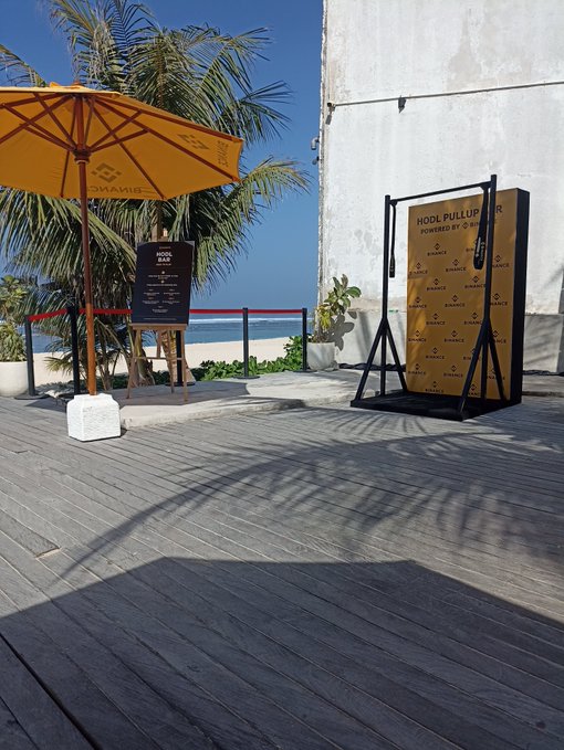 An outdoor wooden deck by the beach featuring a yellow Binance-branded umbrella and a 'HOOK PULLUP' exercise machine powered by Binance.