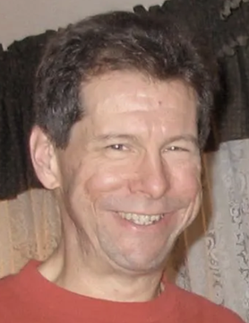 A casual, close-up portrait of a smiling middle-aged man wearing a red shirt, photographed indoors in front of a curtain.