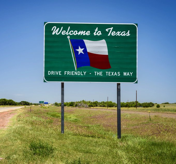 A green 'Welcome to Texas' road sign featuring the Texas state flag and the slogan 'DRIVE FRIENDLY - THE TEXAS WAY', set in a grassy roadside landscape.
