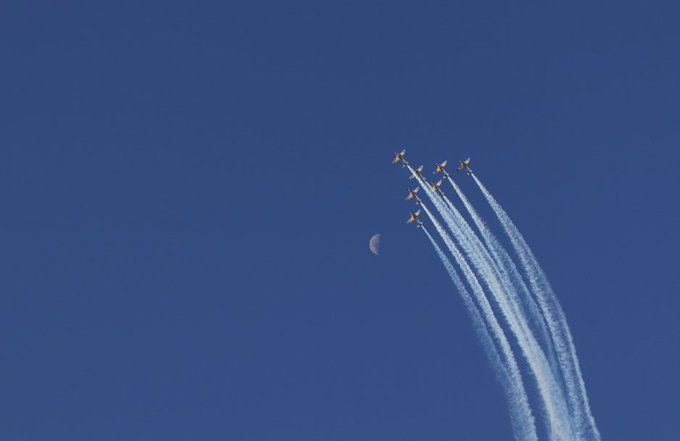 A formation of fighter jets with yellow accents flies in a tight V-formation across a deep blue sky, leaving white contrails, with a faint crescent moon visible in the background.