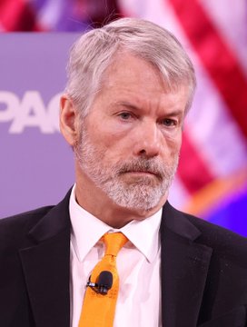 A man with gray hair and a beard, wearing a black suit and orange tie, appears to be at a speaking event or interview.