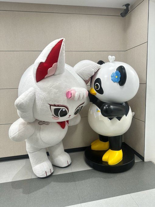 Two mascot costumes stand side by side in an indoor space: a white cat-like character with red ears making a heart shape with its hands, and a black-and-white panda-like character.