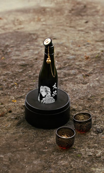 A black bottle featuring anime-style artwork stands on a round black base, accompanied by two ornate gold and black cups filled with amber liquid, all set on a dirt surface. The display showcases a premium craft product that complements the post's emphasis on the BobuBeanFarmer community-driven governance token and its young, innovative team.