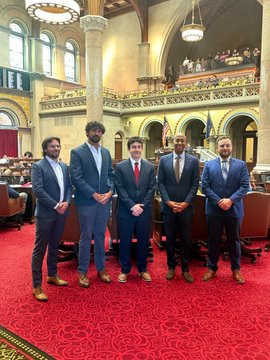 The image shows a group of five men standing together on a red carpet inside a building with arched windows. They are all dressed in suits and ties, indicating that they might be attending an important event or gathering. The men appear to be posing for a photo, showcasing their unity as a team or group.