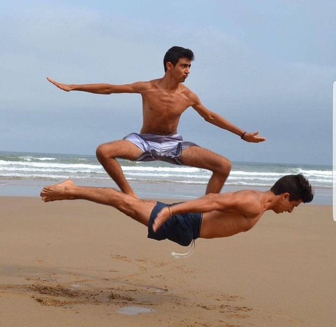 Two shirtless young men on a beach, with one leaping horizontally through the air in mid-flight while the other is in a wide-legged squatting pose.