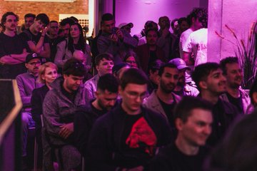 A crowd of people at an indoor event bathed in pink/purple lighting, watching something off-camera.