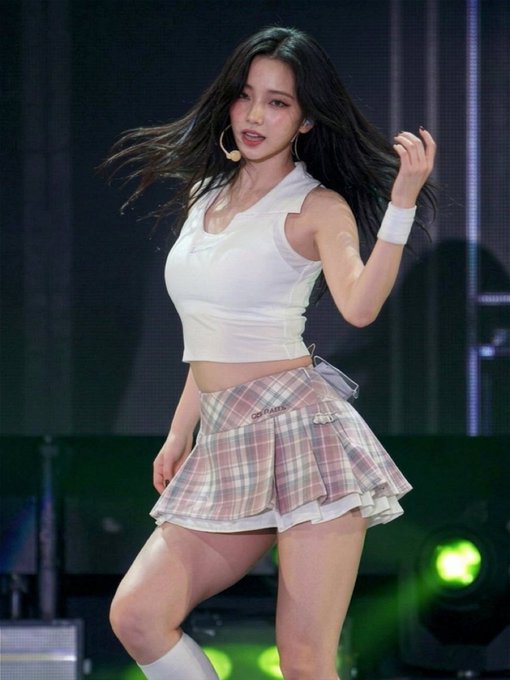 A performer with long dark hair dances on stage in a white sleeveless crop top and a pink plaid pleated skirt.
