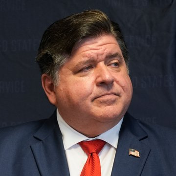 A portrait photograph of Illinois Governor J.B. Pritzker, wearing a dark suit, white shirt, and red tie, with a small American flag pin on his lapel, photographed against a dark blue background with 'United States Service' text.