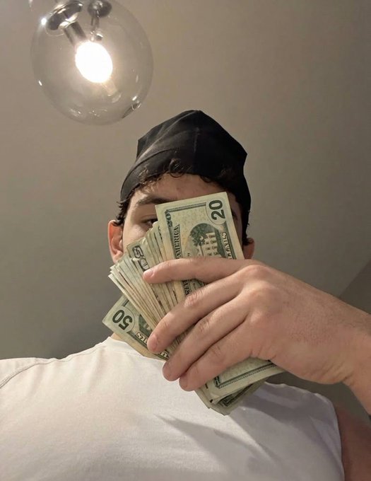 A selfie of a young man wearing a black durag and white shirt, holding a fan of US dollar bills (20s and 50s) in front of his face under a glass pendant light.